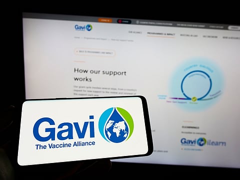 Person holding cellphone with logo of health partnership Gavi, the Vaccine Alliance on screen in front of webpage. Focus on phone display.