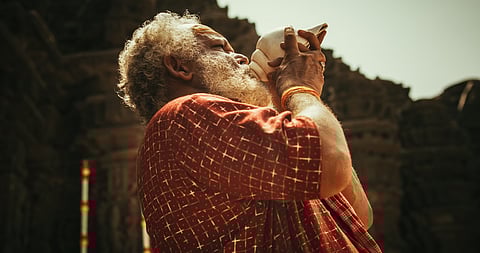 Hinduismo, religión del sur de Asia y metraje cultural: Foto auténtica de un monje indio senior soplando una caracola en un antiguo templo de piedra. Anciano realizando un ritual sagrado en un día de adoración