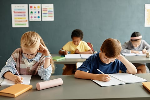 Young studious girl and boy writing in exercise books sitting at desk while studying during lesson at primary school, copy space