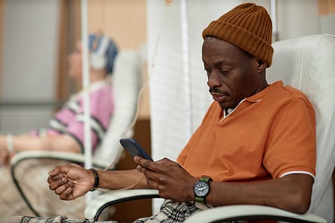 Side view of adult Black man using mobile phone during chemotherapy treatment in clinic copy space
