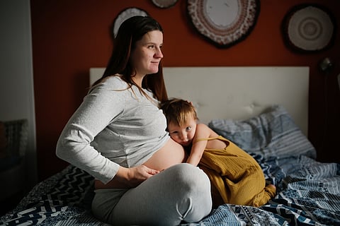 Middle-aged pregnant mother and toddler baby together on bed in bedroom in real interior.