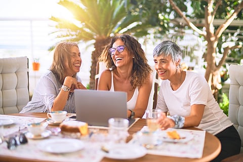 Meeting of middle age women having lunch and drinking coffee. Mature friends smiling happy using laptop at home on a sunny day