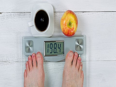 Weight scale, displaying body weight, with bare male feet and whole apple with black coffee in front  on white wooden floor