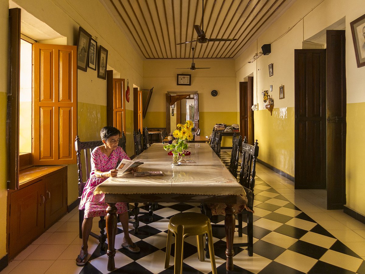 A resident of Lar Santa Margarida sits at the dining table reading the newspaper. The room still has the original chequered flooring and old black-and-white photographs of the Gomes-Pereira family on the walls.