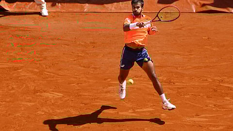 A photo of Sumit Nagal, Indian tennis player, in mid air as he prepares to receive a tennis ball. Sumit is playing on a red clay court and he is wearing an orange tshirt and blue shorts