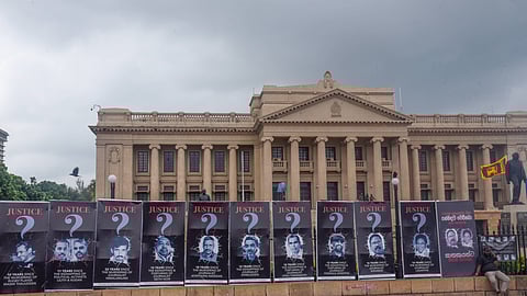 A line of posters showing killings and crimes of impunity during the Rajapaksa government, including crimes against journalists. The poster of Nimalarajan is third from left in a line of posters. His poster has the word 'Justice' on the top and a big gray question mark, a photo of his face, and then a line noting that it has been 22 years since Nimalarajan was murdered. Other cases highlighted include Wasim Thajudeen, abduction of political activists Lalith and Kugan, Keith Noyahr, Prageeth Eknaligoda, Taraki Sivaram, Poddala Jayantha and Lasantha Wickrematunge. You can see the facade of the Presidential Secretariat in the background. 