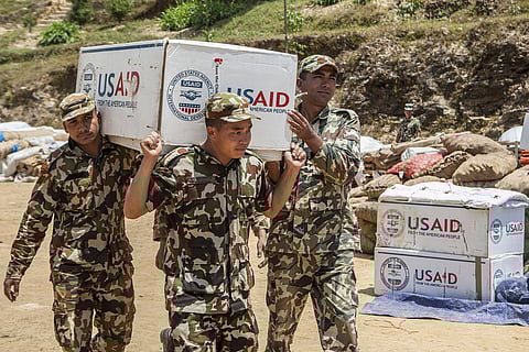 Three Nepali soldiers in camouflage uniforms carry a large box with the USAID logo. 