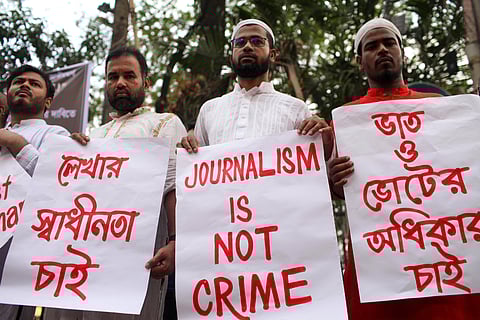 Four men standing in a row holding protest signs in Dhaka. The man in the centre holds a sign that reads 'JOURNALISM IS NOT CRIME' in red letters. The other signs are written in Bengali, with messages calling for freedom of the press. The protest appears to be taking place outdoors, with trees in the background.