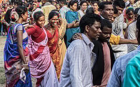 A lively crowd of people from the Oraon community in Purnea celebrates the spring festival of Sarhul. Men and women link arms and dance in traditional attire, with women in colourful sarees smiling and moving energetically, while others watch in the background. The atmosphere is festive and joyful.