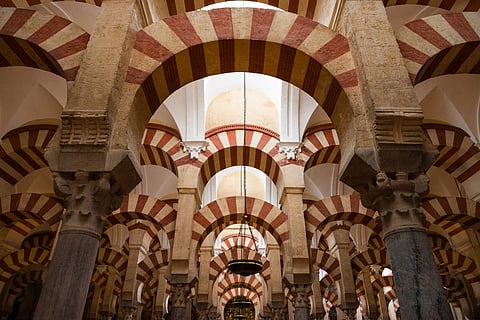 Interior view of the Mosque-Cathedral of Córdoba in Spain, showcasing a series of red and cream striped horseshoe arches supported by columns. The architectural design features intricate Islamic and Gothic elements, with a hanging chandelier in the centre. 