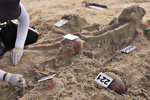An archaeologist at the the Chemmani mass grave site excavates partially unearthed human skeletons from the sandy ground, with numbered evidence tags placed near the bones.