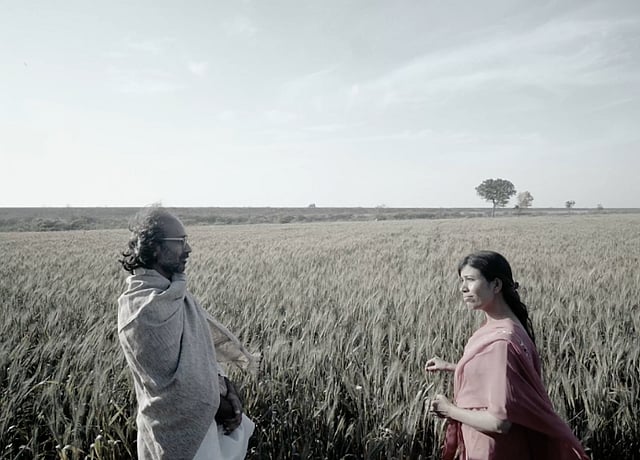 A spectacled farmer on the left, and a poet on the right, stand in front of a field. The photo is in greyscale. This is a still from Indus Echoes, a film shot in Sindh, Pakistan