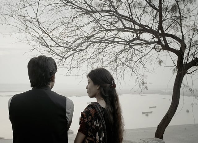 A man and a woman stand next to each other. The man is looking out at a lake while the woman is looking at him. A tree is in the right corner. The photo is in greyscale. This is a still from the movie Indus Echoes, shot in Sindh province, Pakistan.