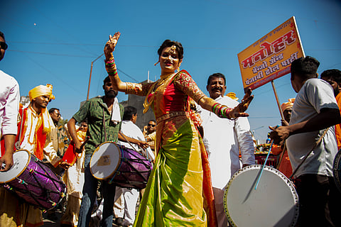 A member of the hijra community dances at a religious procession in Maharashtra, wearing a bright green and red sari. Drummers surround them and celebrate in the street. A yellow signboard with Marathi text is visible in the background under a clear blue sky.