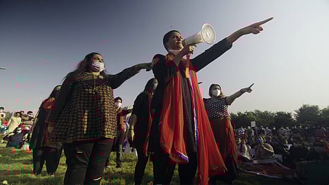 A still from This Stained Dawn depicts a group of women wearing black and red in formation at Aurat March. Behind them, a mostly female audience sits on the grass and watches the performance.