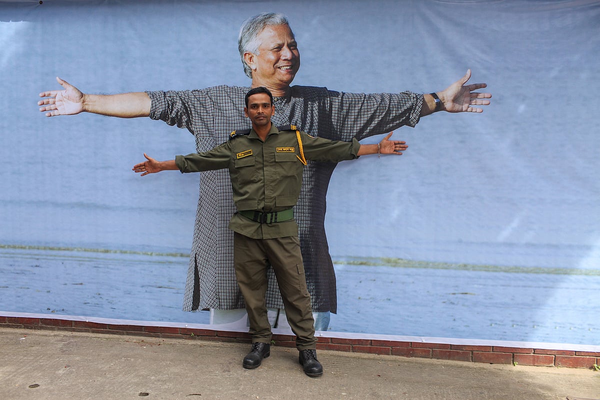A man in a green security guard uniform stands with arms spread out in front of a poster of Muhammad Yunus in a grey checked 