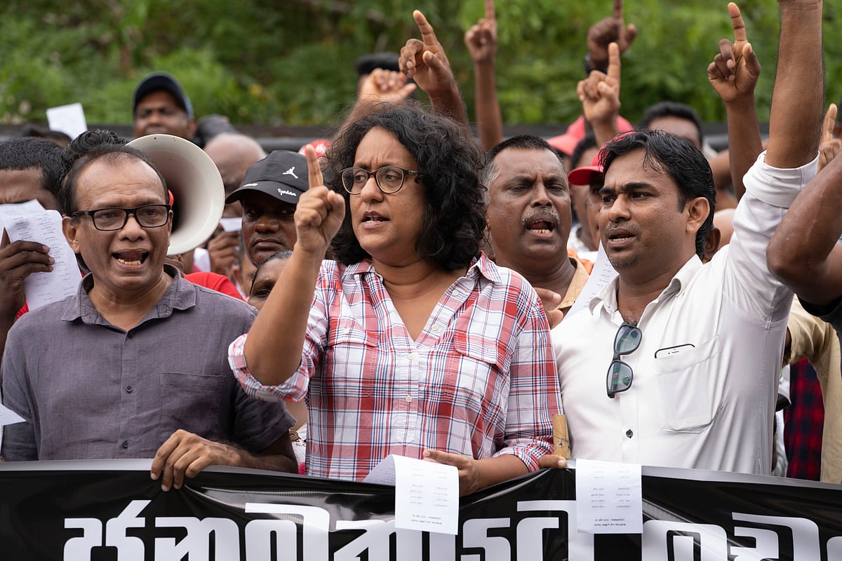 Harini Amarasuriya dressed in a red and white checked shirt standing behind a black banner with white lettering held by two m