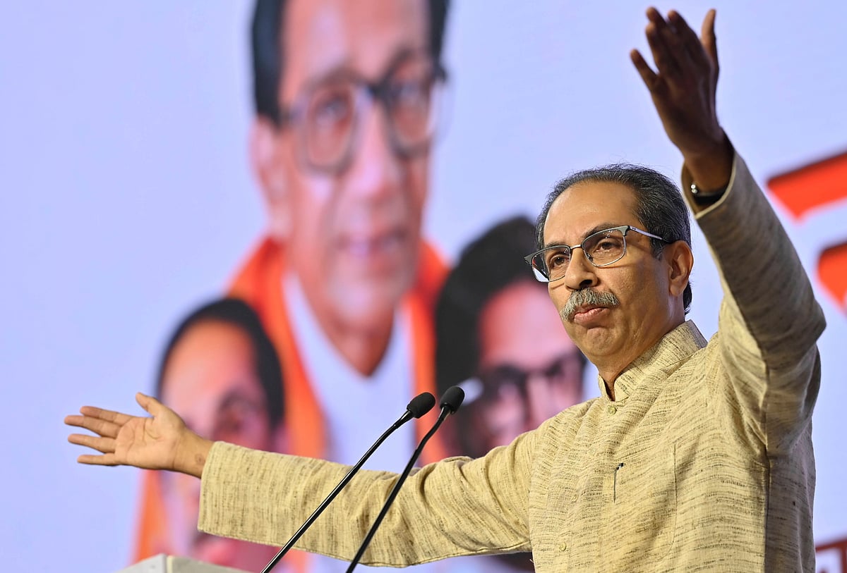 Maharashtra politician Uddhav Thackeray, wearing a beige kurta, stands in front of a mike with his arms spread out. In the ba