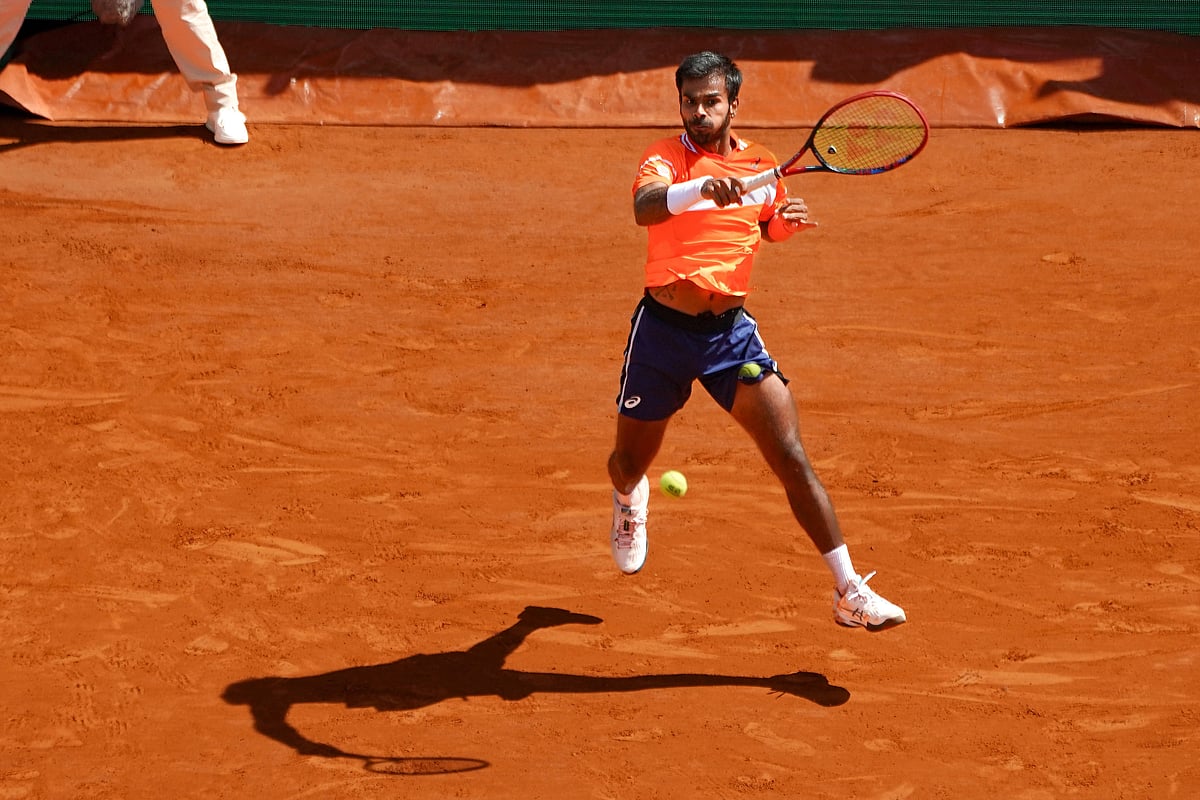 A photo of Sumit Nagal, Indian tennis player, in mid air as he prepares to receive a tennis ball. Sumit is playing on a red c