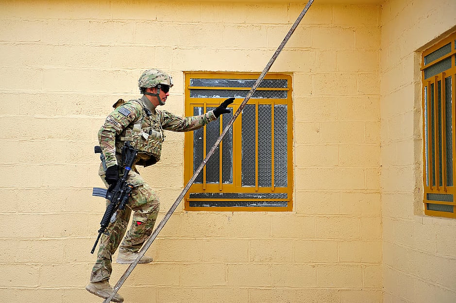 US Army National Guard 1st Lieutenant Ronald Snyder, part of the Provincial Reconstruction Team in Farah, Afghanistan in 2012. Photo shows Snyder climbing a ladder outside a house with pale yellow walls. There is a window in the middle of the wall with glass panes.  There is another window on the right hand side of the photo