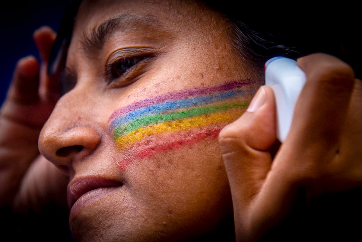 A close up of a person's face. There is a pair of hands visible in the photo drawing on the pride colours of Purple, blue, green, orange, yellow and red with a white pen. 