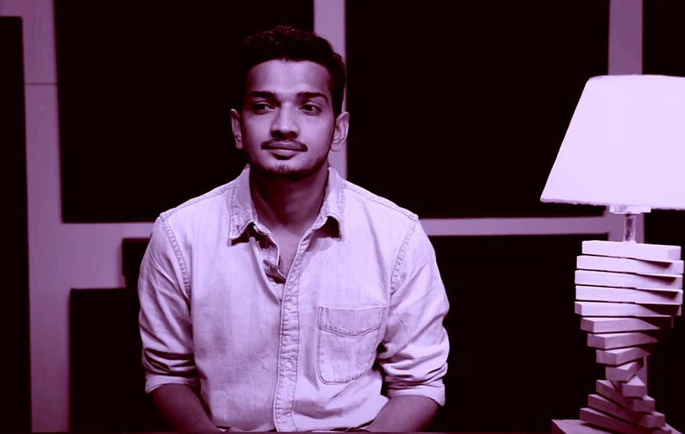 Photo of a man sitting in a room, next to a lamp. There is a pink filter over the photo for some reason. This is a screengrab from a video shot by comedian Munawar Faruqui titled 'Munawar Faruqui leaving comedy'. Faruqui was arrested before a comedy gig by a group of Hindu vigilantes who thought his jokes could violate the Penal Code and other laws - even though his gig had not started yet. 