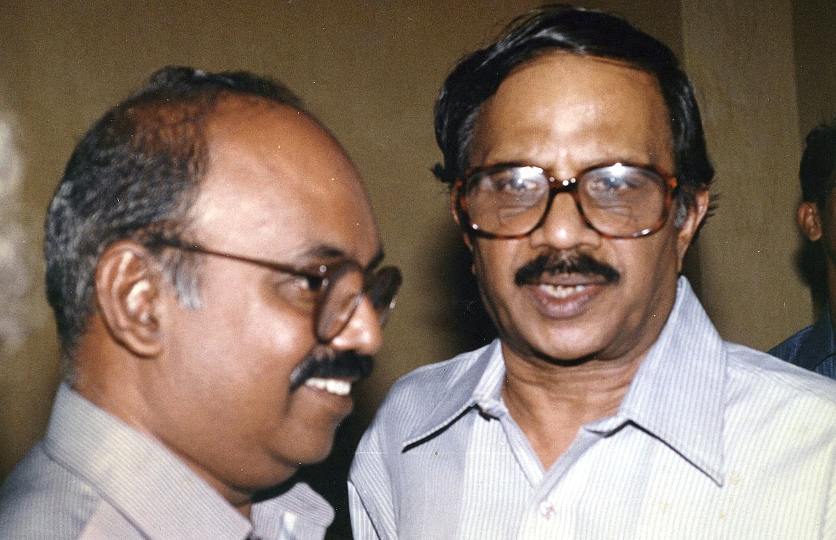 An old photograph of M T Vasudevan Nair (right), wearing glasses and a light striped shirt, smiling while facing the camera. 