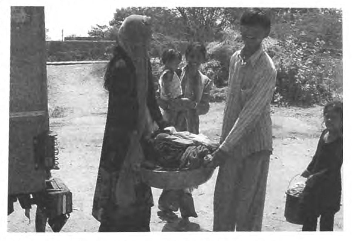 Two people carrying a basket of laundry with a woman holding a kid in her arm in the back. A child carrying a bucket in the c