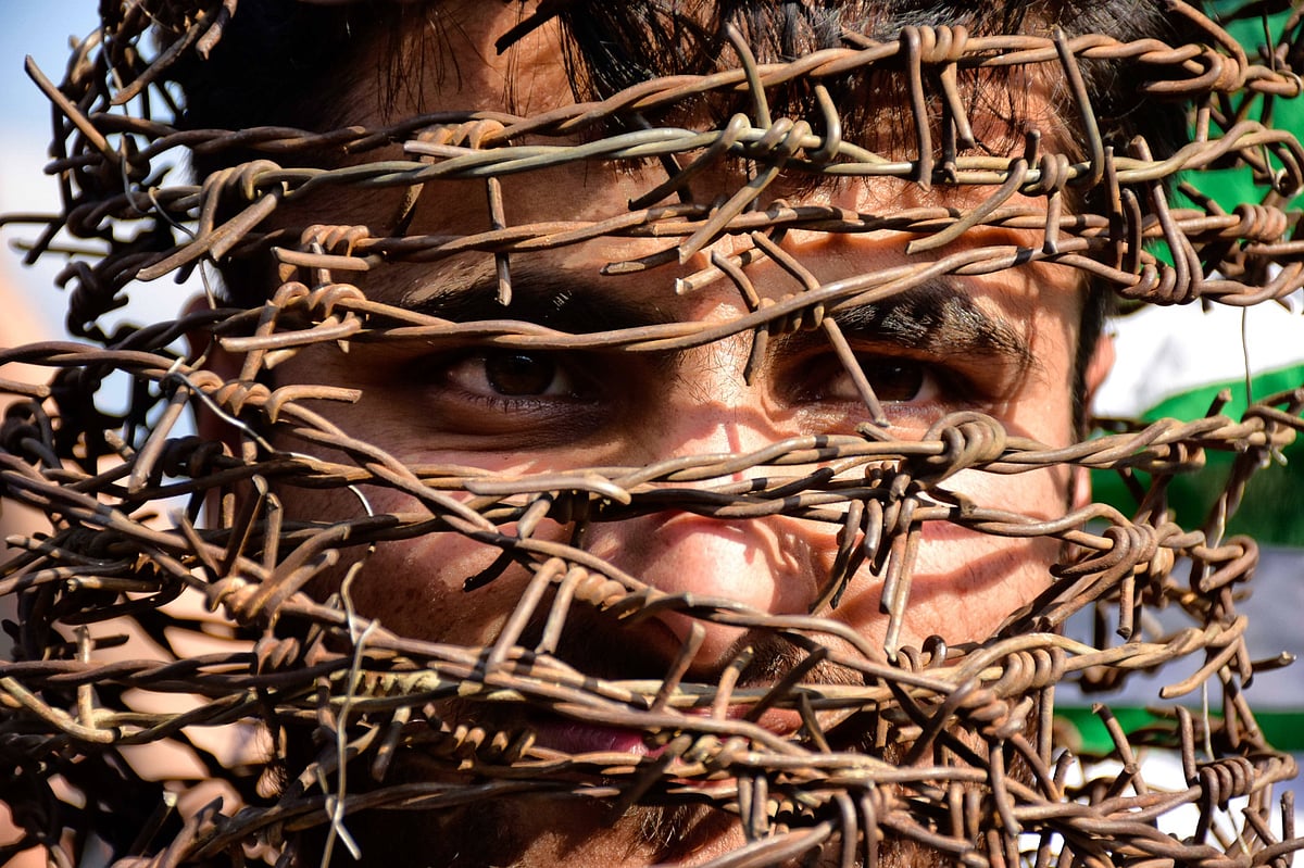 A close-up photograph of a man's face partially obscured by tangled and rusted barbed wire as he stare through the gaps. The 