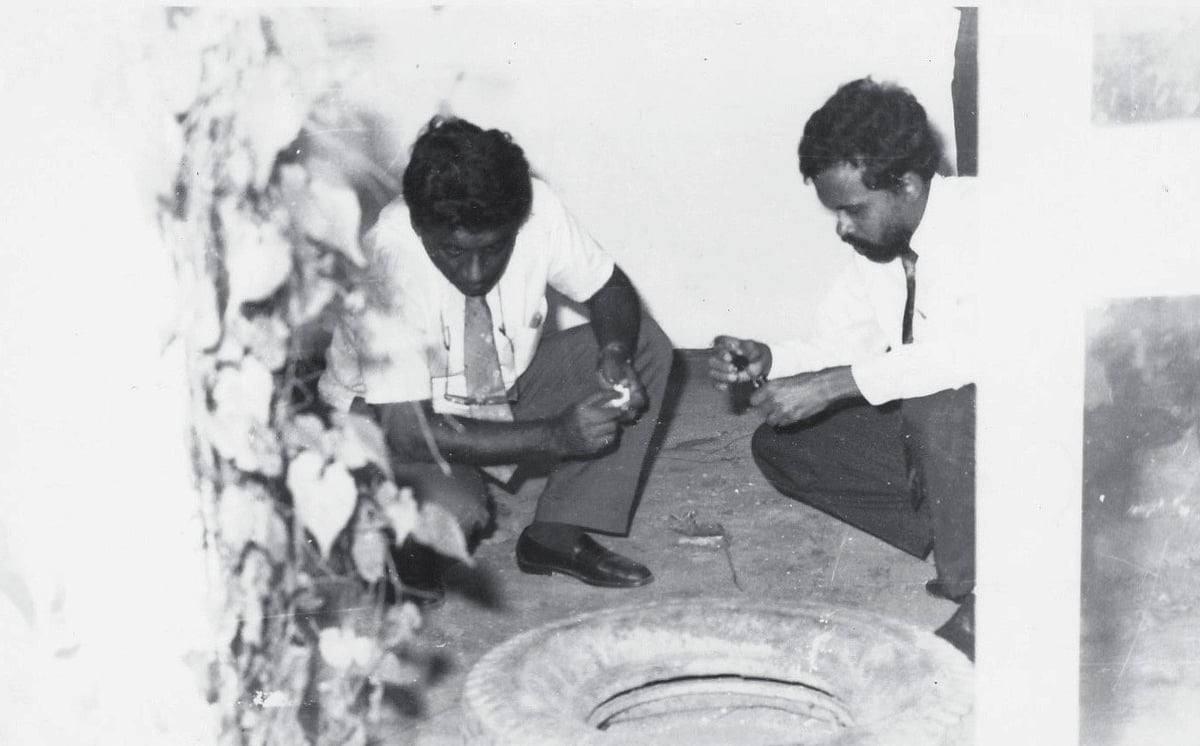 An old black and white photograph of two men in white shirts and ties crouching on the ground near a tyre. The man on the rig