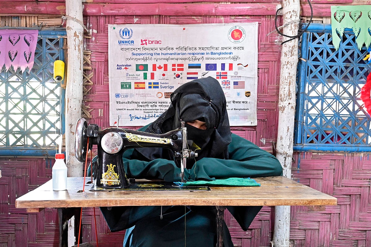 A Rohingya woman dressed in an abaya and hijab works at a sewing machine in a. refugee camp. A banner with logos of donor org
