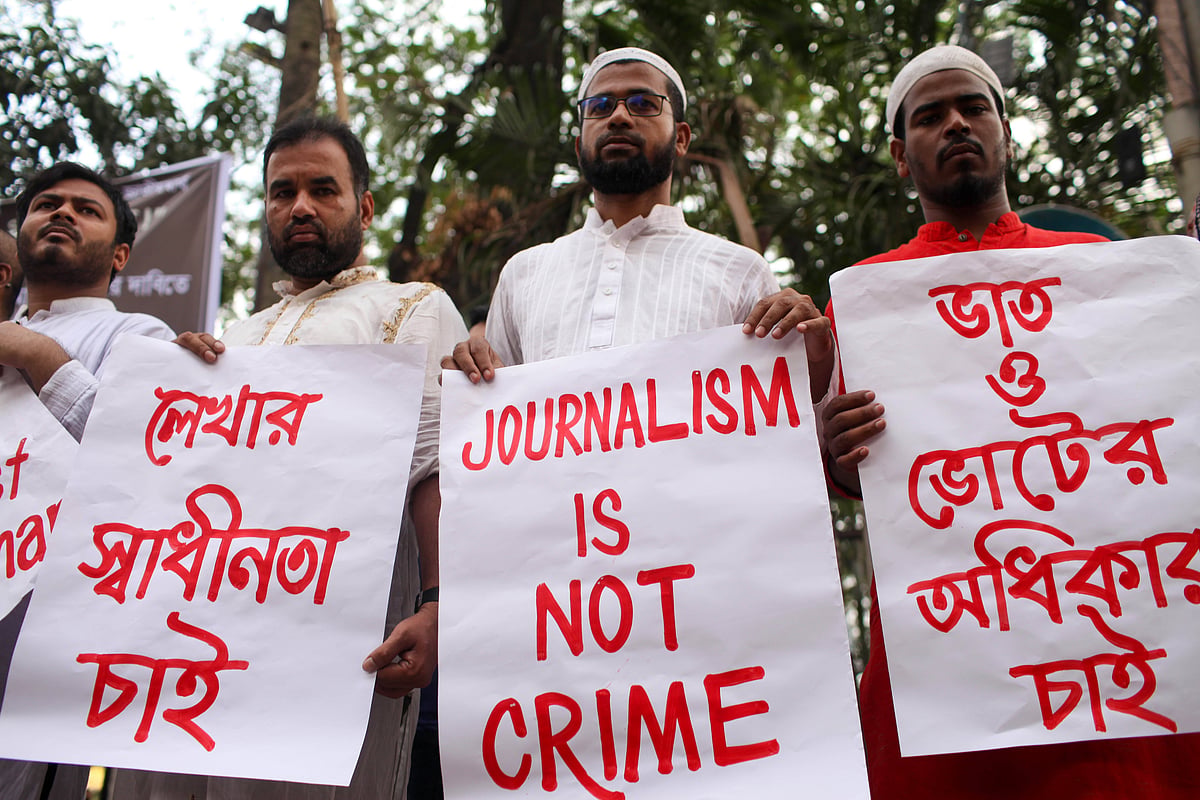 Four men standing in a row holding protest signs in Dhaka. The man in the centre holds a sign that reads 'JOURNALISM IS NOT C