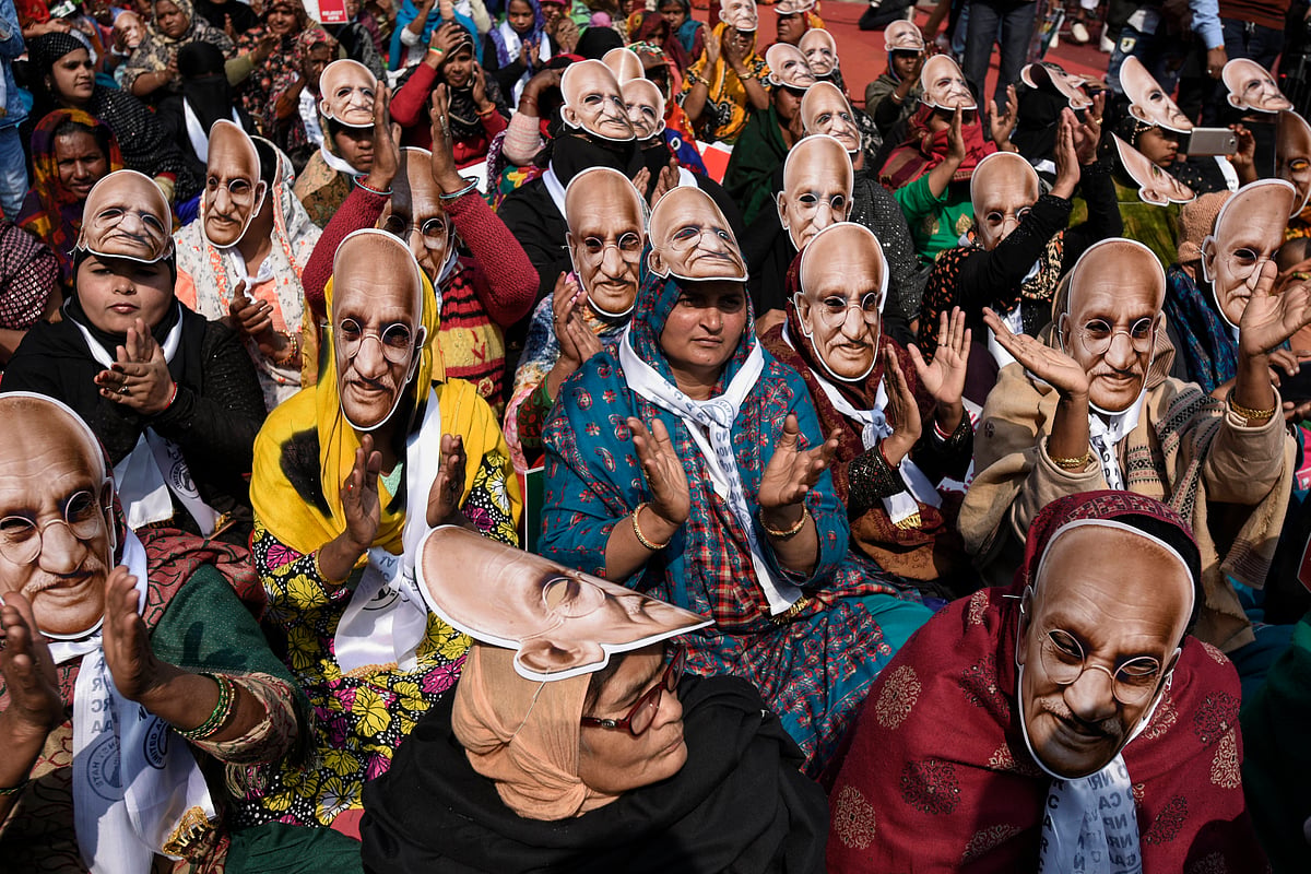 A large group of women are sitting together, wearing masks of Mohandas Karamchand Gandhi's face, participating in a peaceful 