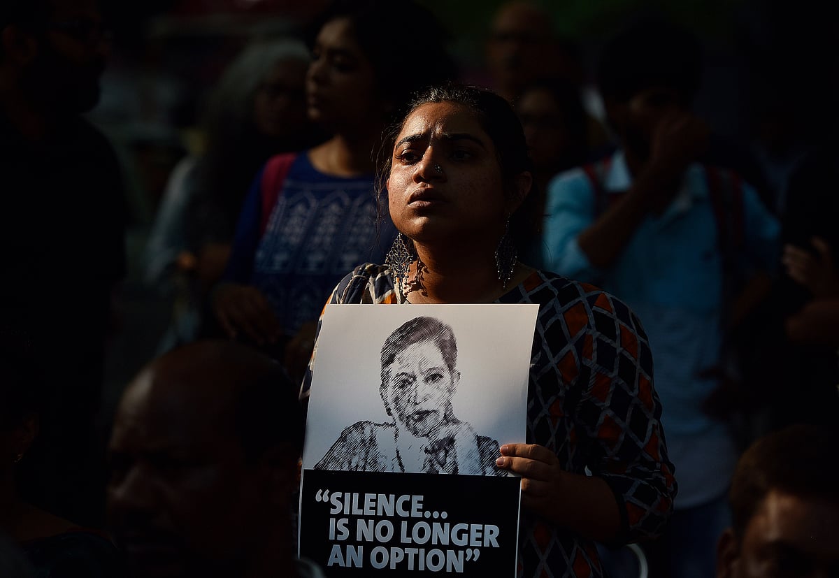 A woman stands in a crowd holding a black-and-white poster featuring a portrait of Gauri Lankesh and the words 'SILENCE... IS