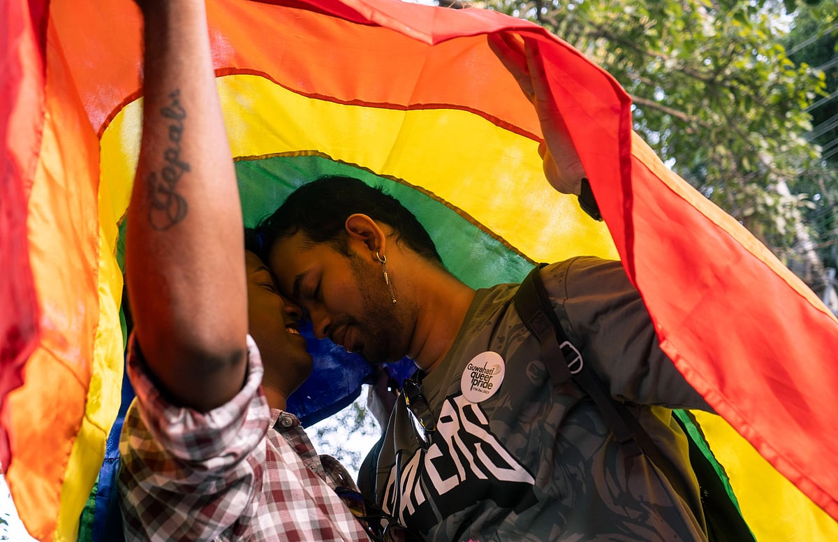 Two men stand face to face under a rainbow pride flag at a Pride Parade in Guwahati, Assam. One of them wears a plaid shirt w
