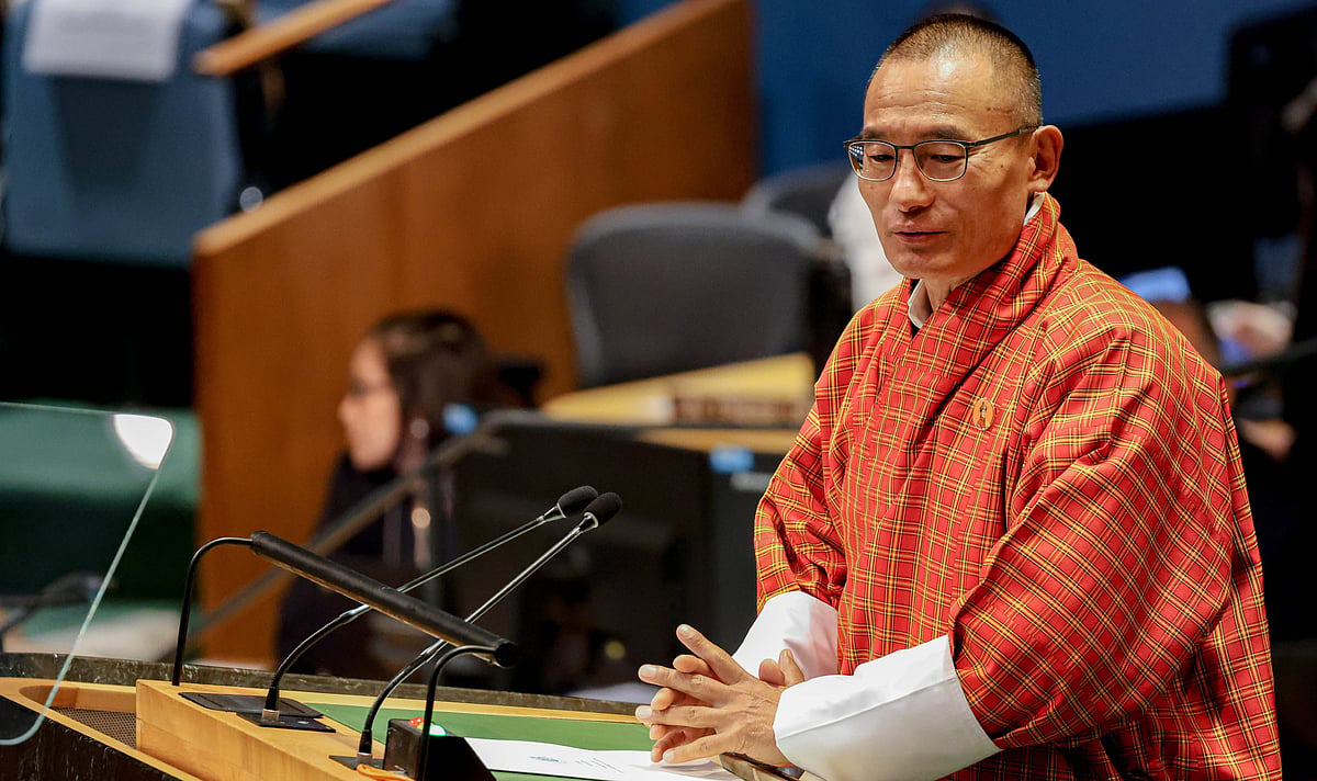 Tshering Tobgay in traditional Bhutanese attire, wearing glasses and a red patterned gho, stands at a podium delivering a spe