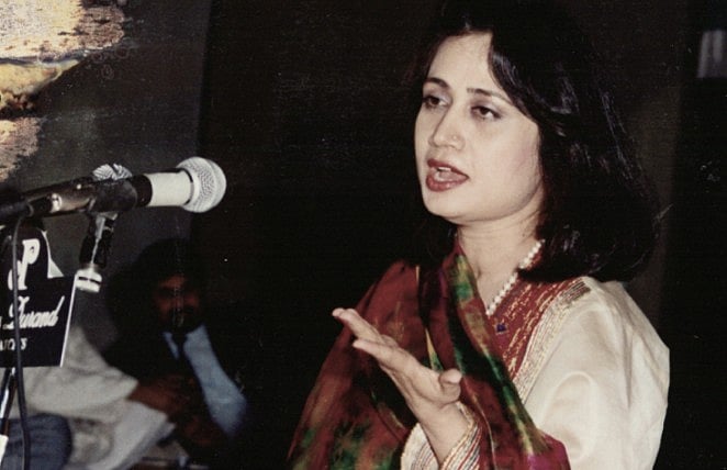 A photograph of Parveen Shakir in speaking into a microphone at a public event. She is mid-sentence, with one hand raised in 