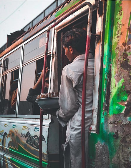 Photo of a man leaning out of a bus. He is holding a bowl of food, wrapped in bundles. The other passengers are looking out of the window. 