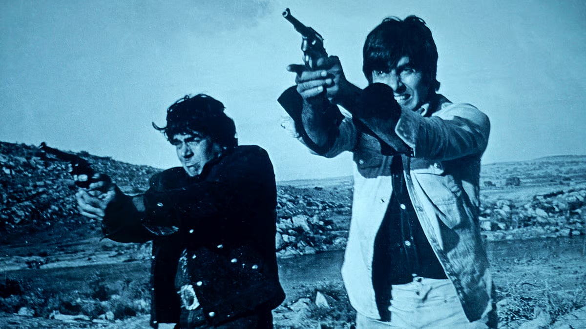 A still from the Hindi blockbuster Sholay, starring Dharmendra (left) and Amitabh Bachchan (right), both holding guns with a hilly landscape in the background