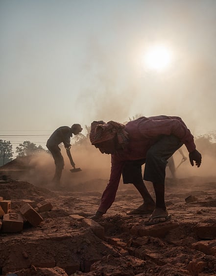 Photo of workers in a brick factory. Man in the foreground with his head covered, laying a brick. There is another man holding an implement in the background. The air is filled with fine red dust. 