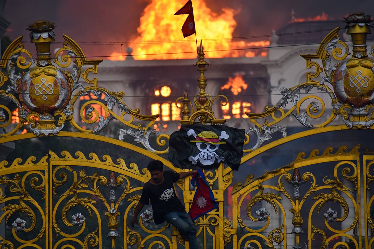 A man hanging from an ornate gate with a pirate flag hung on it as a white neo-classical building burns in the background.