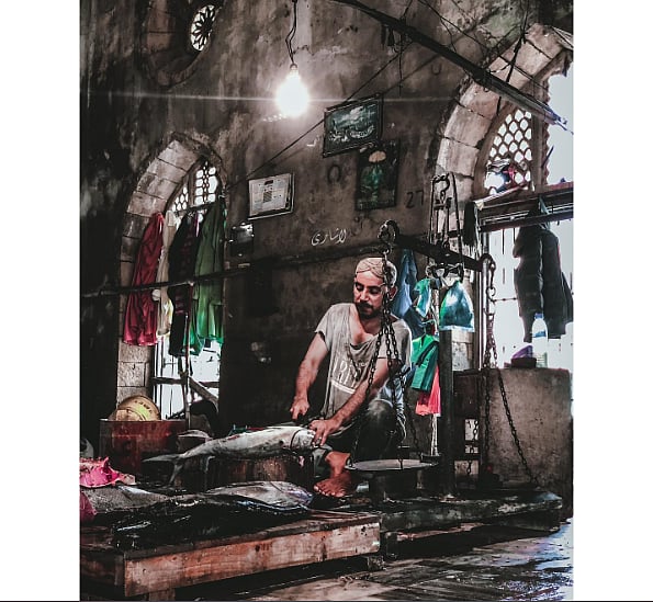 Photo of a man in a fish market, cutting fish. The walls are gray and there are large windows in the shape of a minaret in the background. 