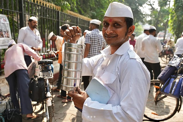 Photo of a dhabbawala (man who delivers tiffin boxes) smiling and holding up a tiffin box. 