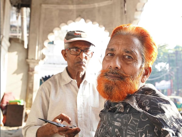 Photo of a man getting his beard trimmed. His hair and beard are dyed red, though his eyebrows are dark. 