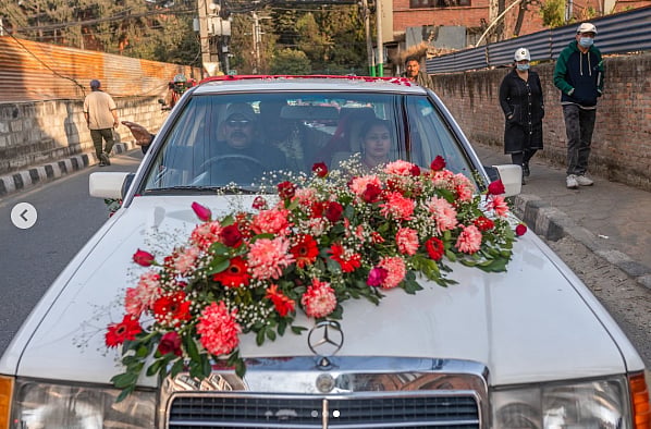 Photo of a car covered in flowers. A man and woman sit in the front seat. A bride garlanded in flowers can be seen faintly in the back seat. On either side of the pavement, people are walking past. The walls are brick, topped with asbestos sheets. 