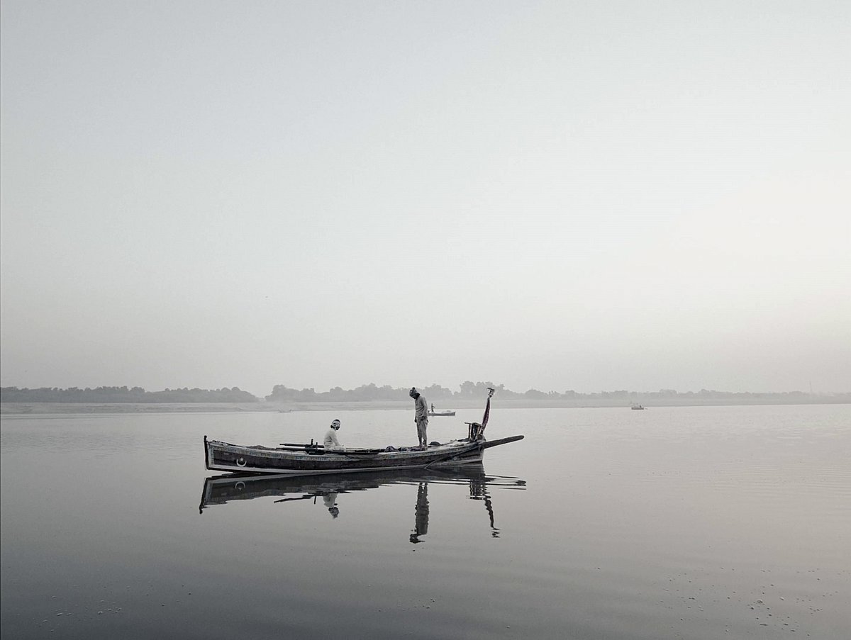 Two men on a boat floating on a still lake. Photo is in grey scale. This is a photo from Indus Echoes, a film shot in Sindh, 