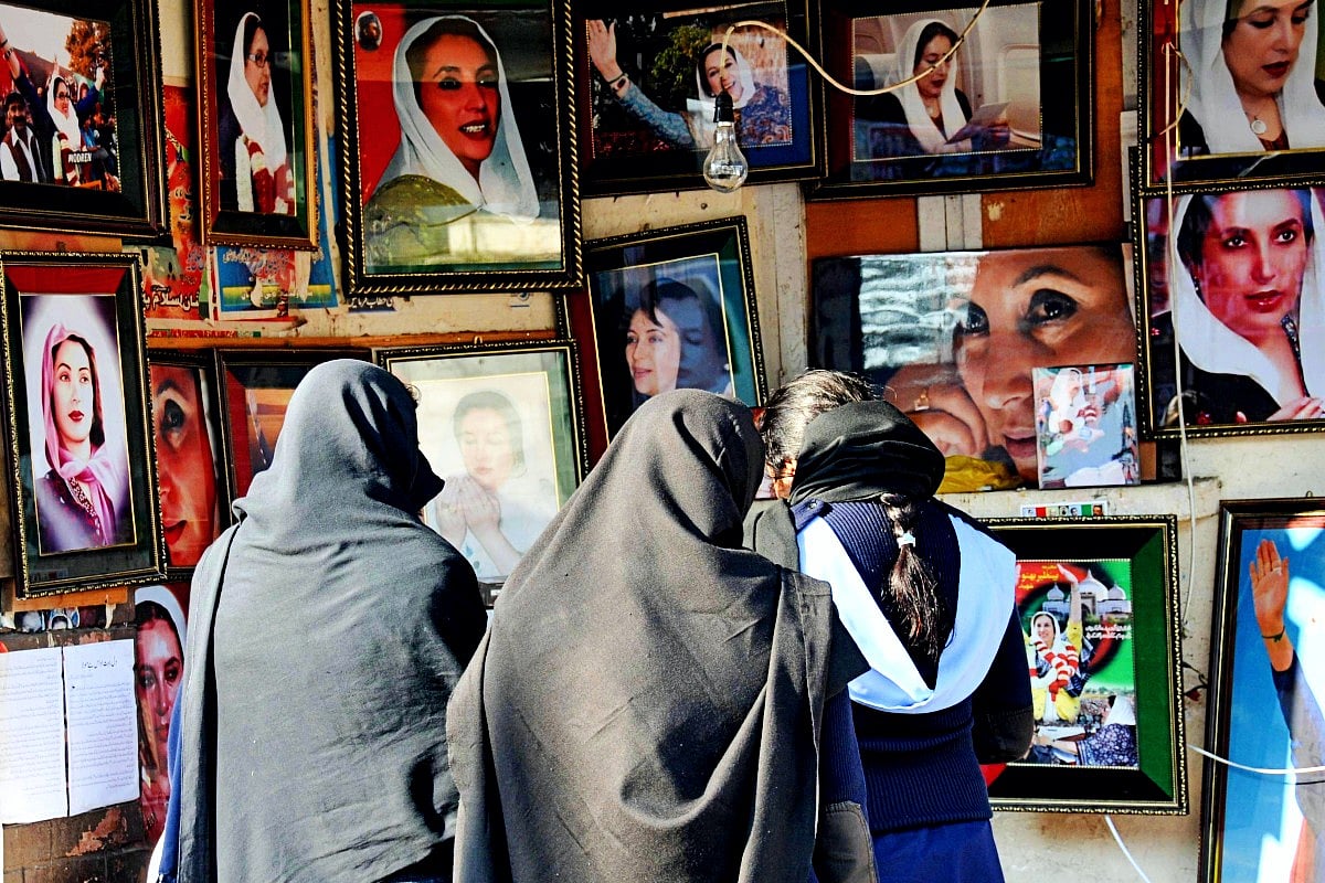 A group of women, seen from behind and wearing dark headscarves and shawls, stand closely together in front of a display wall