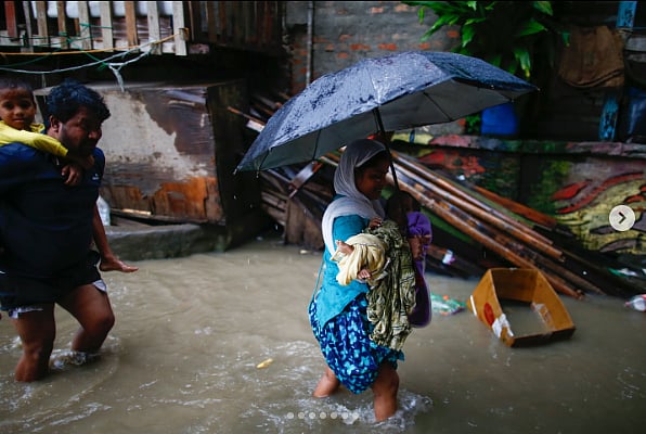 Photo of a woman wading through floodwater carrying a young child and holding an umbrella. She wears a white shawl covering her head and a blue shalwar top, and flowered blue pants. Behind her a man is carrying another young child on his back. He is wearing a dark blue tshirt and shorts.
