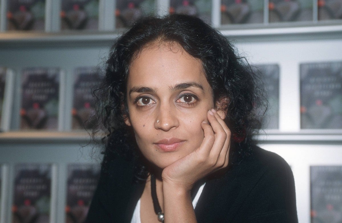Arundhati Roy rests her chin on her hand and looks directly at the camera. She is seated indoors in front of shelves filled w