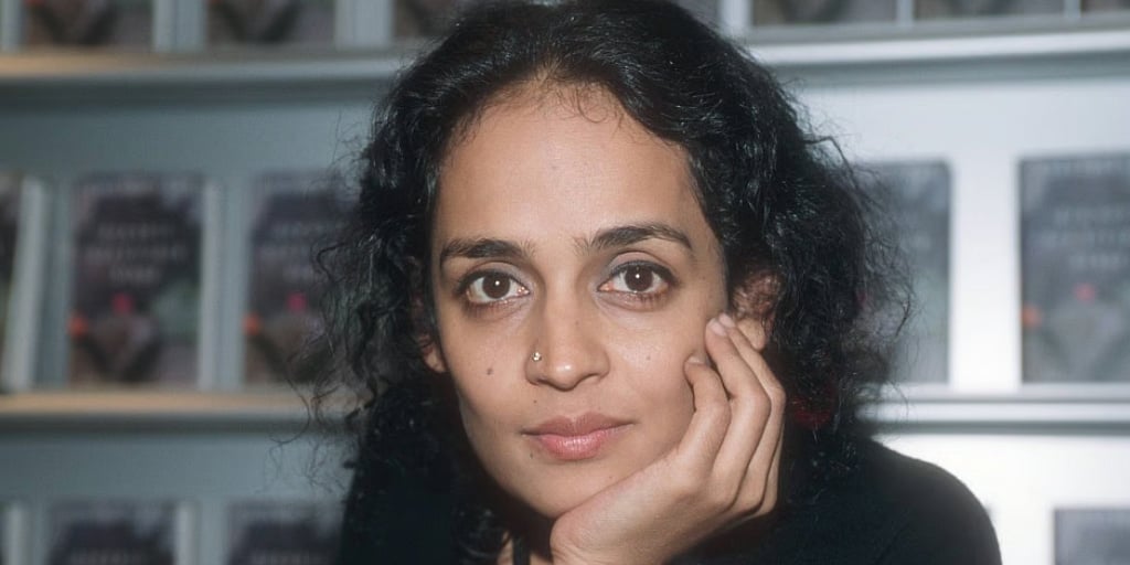 Photo of Arundhati Roy looking into the camera. The covers of her books can be seen faintly in the background.